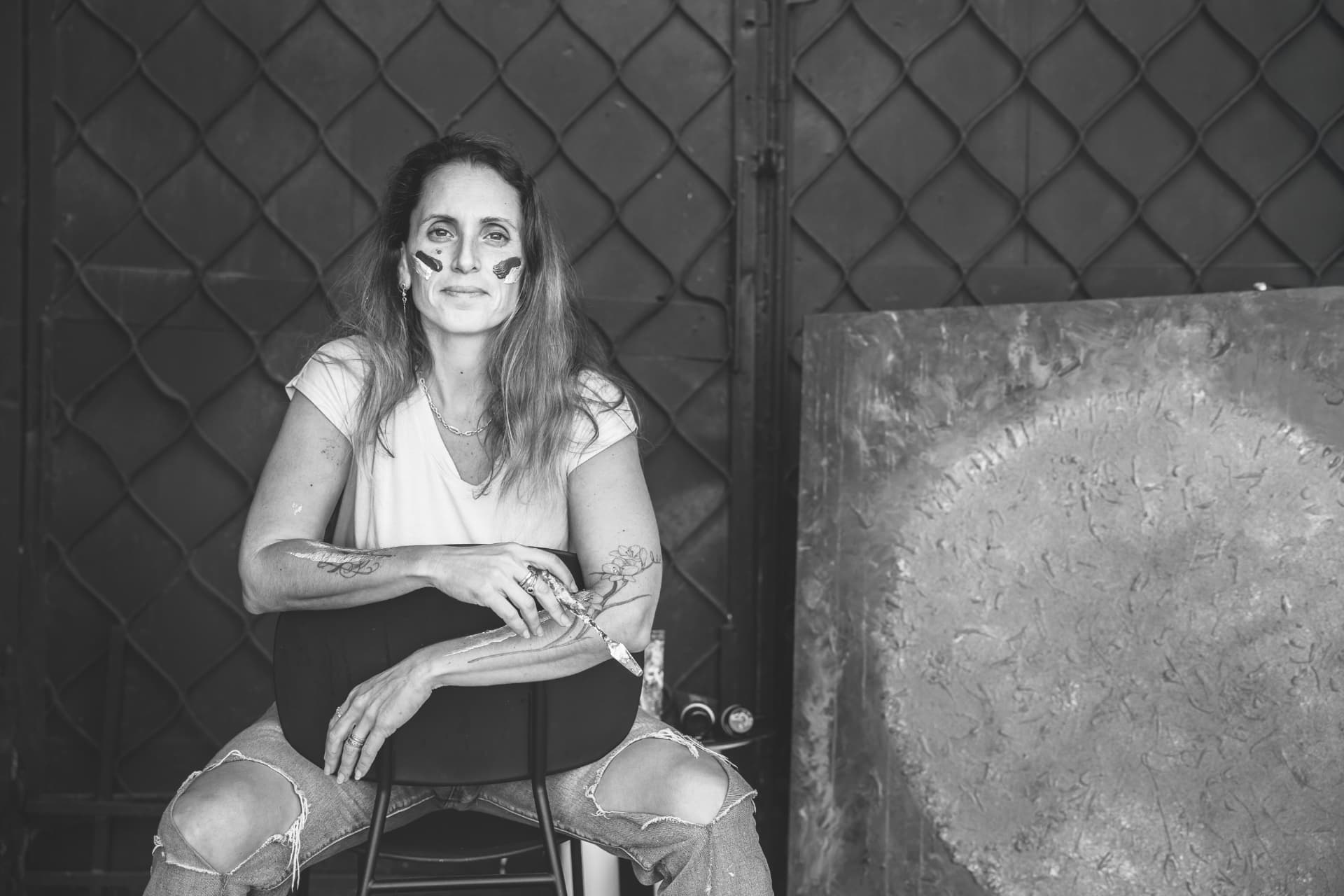 Nili Stein in her studio with paint on her face and hands, holding a painting tool